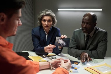 İki dedektif bir şüpheliyi sorguluyor. Masada parlak ışık altında kanıt sunuyorlar. İki dedektif de şüpheliyi dinlerken, kelepçelerken ve turuncu tulum giyerken odaklanmış durumdalar.