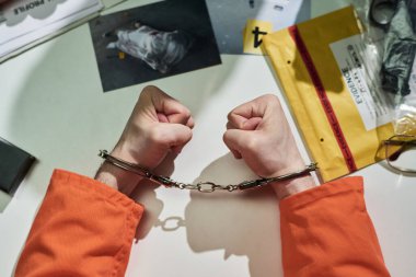 Elleri kelepçeli, turuncu tulumlu, kanıt zarfları, fotoğraflar, belgeler ve kelepçe ile masanın üzerinde dinlenen eller soruşturmacı bir atmosfer yaratıyor.