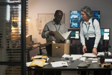 Çalışma arkadaşları, ortak ofiste kağıt ve klasörlerle kaplı masalar, arka planda veri görüntüleyen ekranlar ve işbirlikçi çalışmalar içeren önemli belgelere odaklandılar.