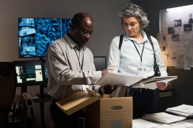 Dedektifler bir ofiste rozet takıp dava belgelerini inceliyorlar. Etrafları çeşitli teknoloji ve kağıt parçalarıyla çevrili.