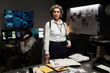 Dedektifler modern tesisteki suç mahalli belgelerini inceliyor. Etrafında birden fazla monitör ve dosya var. İş arkadaşı arka planda ayrıntıları tartışıyor