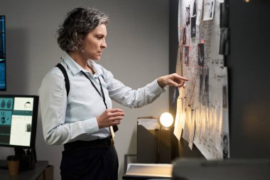 Kısa saçlı bayan dedektif, ofis ortamında olay yeri kanıtlarını ilan panosunda inceliyor.