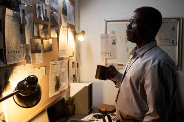 Masasında duran Afrikalı Amerikalı bir adam elinde bir kupa ve masasının üzerinde bulunan ve masa lambasıyla aydınlatılan çeşitli belgelerle dolu inceleme tahtası.
