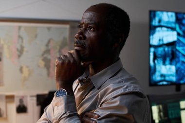 Modern ofis ortamındaki verileri düşünceli bir şekilde analiz eden bir adam büyük bir harita ve bilgi içeren çoklu ekran düzeneği sunar