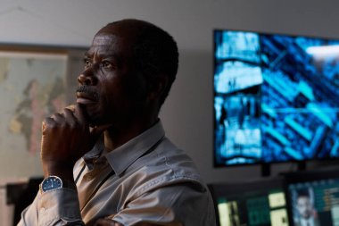 Modern ofiste derin düşünen yaşlı Afrikalı Amerikalı bir adam ileri teknoloji ve veri tasvir eden büyük ekranlarla dolu. Çene üzerinde el tefekkür ve analizi gösterir