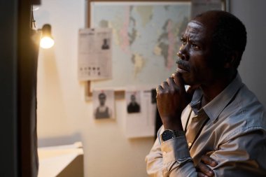 Ofiste duran yaşlı adam duvardaki dünya haritasına özenle bakıyor. Etrafında masa lambasıyla aydınlatılmış fotoğraflar ve belgeler var.