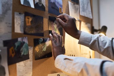 Belirsiz aydınlatılmış odadaki ilan tahtasına resim ve notlar iliştiren eller, soruşturma altındaki suç mahalliyle ilgili çeşitli kanıtlarla birlikte.
