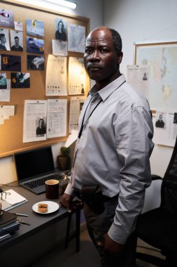 Yaşlı Afro-Amerikan bir adam ofis masasında duruyor, olay yeri fotoğraflarını ve belgelerini inceliyor, ciddi bir ifadeyle, kameraya bakıyor.