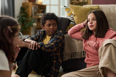 Bir grup çocuk kanepede rahatça oturup sohbet ediyor ve gündelik zamanlarını birlikte geçiriyorlar. Pembe kazaklı çocuk, dizliğine yaslanıyor.