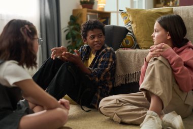 Üç çocuk oturma odasının rahat zemininde, dekoratif yastıklar ve yumuşak ışıklandırmayla neşeli bir sohbete dalıyorlar. Sıcak bir ortam yaratıyorlar.