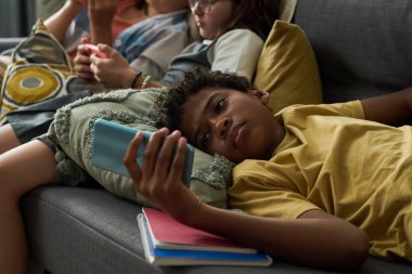 Kanepede uzanmış telefonu tutan genç bir çocuk, odaklanmış ve nişanlı görünüyor. Arka planda cihazları olan diğer bireyler de var. Rahat ve rahat bir ortam yaratıyorlar.