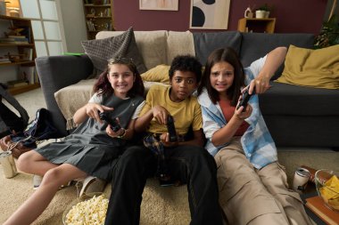 Üç çocuk kanepede oturmuş video oyunu oynuyorlar ellerinde kontrolörlerle ve yerde patlamış mısırla rahat oturma odasında canlı bir atmosfer yaratıyorlar.