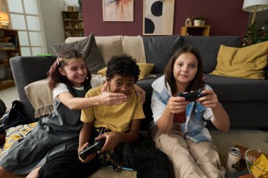 Üç arkadaş oturmuş, modern dekorları, kitap rafları ve yumuşak ışıklandırmasıyla rahat oturma odasında video oyunlarının keyfini çıkarıyorlar. Sıcak bir ortam yaratıyorlar.