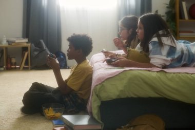 Yatak odasındaki kanepede video oyunu oynayan bir grup çocuk ve çeşitli kişisel eşyalar etrafa saçılmış. Rahatlamış atmosfer ve paylaşılan aktivitelere odaklan