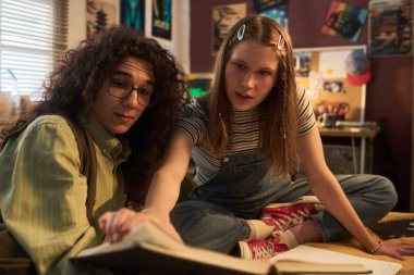 İki genç yurt içinde birlikte okuyorlar, kitaba odaklanıyorlar. Sıcak ışıklandırma arka planda görünen posterler ve dekorasyonlarla rahat odayı aydınlatır