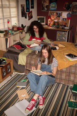 İki kız halı döşemede ve yatakta iyi dekore edilmiş retro yatak odasında oturuyor, biri gitar çalarken diğeri rahat ve üretken bir atmosfer yaratıyor.