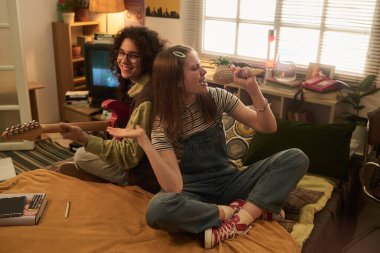 Evde müzik dinlemekten hoşlanan iki arkadaş, biri gitar çalıyor ve diğeri mikrofona tutkuyla şarkı söylüyor, neşeli ve neşeli bir atmosfer yaratıyor.