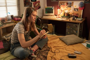 Genç bir kadın yatakta oturmuş, müzik dinlerken deftere bir şeyler yazıyor. Arka planda kişisel retro eşyalar ve bilgisayar masalarıyla dolu bir oda nostaljik bir his yaratıyor.