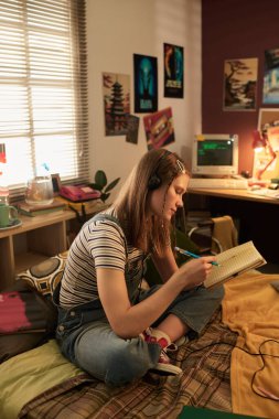 Genç bir kadın rahat yatak odasında kulaklıklar takmış, bağdaş kurmuş, açık bir kitap tutuyor, duvarda posterler ve retro dekorasyonlarla çevrili, masada bilgisayarla oturuyor.