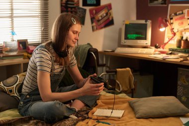 Genç bir kadın, yatak odasında elinde eski model bir bilgisayarla oturmuş, elinde bir bardak tutuyordu. Etraf kitaplık ve çeşitli retro ögelerle çevrili, rahat atmosfer yaratılıyor