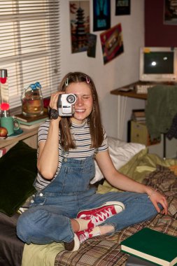 Yatakta oturmuş gülümseyen bir genç, eski bir kamerayı tutuyor, anları yakalamaktan zevk alıyor. Yatak odası çeşitli eski eşyalarla dolu, nostaljik bir atmosfer yaratıyor.