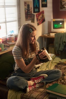 Genç bir kadın, elinde antika kamerayla rahat bir odada oturuyor. Posterler ve antika eşyalarla çevrili. Günlük kıyafetleri ve rahat duruşuyla nostaljiyi çağrıştırıyor.