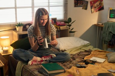 Rahat yatak odasında ders kitapları, aletler ve okul malzemeleriyle çevrili bir kızı inceliyor, pencerenin yanındaki derslerine panjurlarla odaklanıyor, sıradan bir anı yakalıyor.