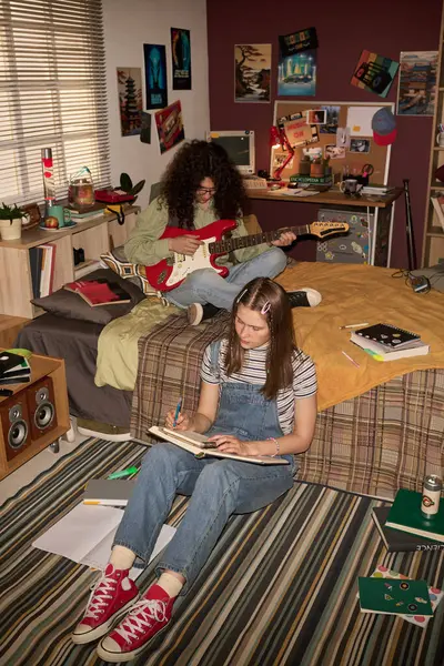 İki genç yatak odasında posterler ve retro çalışma malzemeleriyle etkinlik yapıyor. Bir genç kitap okurken diğeri yatakta kırmızı elektro gitar çalıyor.