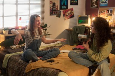 İki genç arkadaş, biri yatakta, diğeri yerde oturuyor, gülümsüyor ve sohbet ediyorlar rahat ve süslü bir retro yatak odası düzeninde.