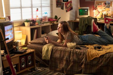 Oyun kumandanıyla yatakta oturan, etrafı kitaplar ve retro dekorlarla çevrili biri. Eski monitör nostaljik dokunuş ekleyerek bir oyun gösteriyor