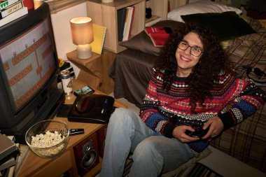 Kıvırcık saçlı, gülümseyen ve kitap rafları ve atıştırmalıklarla dolu bir odada retro video oyunu oynayan biri. Sıcak ışıklandırma ve rahat oturma odasıyla dekore edilmiş.