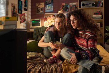 İki arkadaş, oturma odasında eski bir film izliyorlar. Eski moda dekorlar ve antika bilgisayar ekranı da dahil olmak üzere duvarlarda eski moda eşyalarla dolu posterler var.