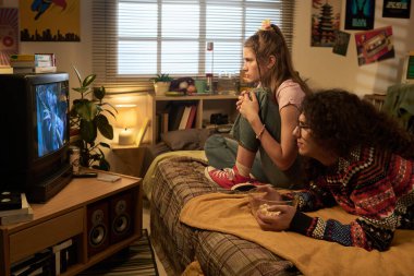 Oturma odasında rahat bir kanepede oturan iki arkadaş ev dekoru ve ortam aydınlatmasıyla çevrili televizyon izlerken sıcak ve rahat bir atmosfer yaratıyor.