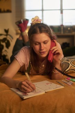 Masasında bir defter ve arka planda iç mekan bitkileriyle rahat, sıcak aydınlatılmış bir odada notlar yazarken genç bir kadın telefon görüşmesi yapıyor.