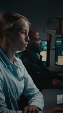 Beyaz kadın güvenlik görevlisinin CCTV ekranını izlerken orta dikey görüntüsü, şüpheli bir olay fark ediyor, Afrikalı Amerikalı erkek ortağa haber veriyor, o da telsizden devriyeye bildiriyor.