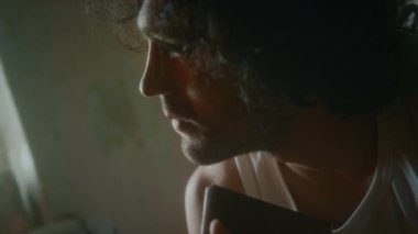 Kirli sakallı, fanilalı, elinde not defteriyle oturan, umursamaz ve başını eğmiş, hayatının anlamını sorgulayan yorgun ve depresif bir adamın yakın çekimi.