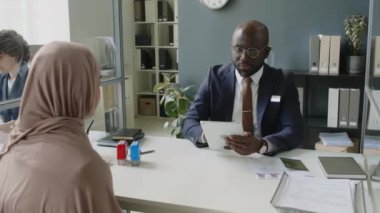 Dijital tabletle kendinden emin Afrikalı Amerikalı erkek vize memurunun tesettürlü Müslüman kadınla röportaj yaptığı orta boy bir fotoğraf, elçilikte planlanan iş gezisi hakkında sorular soruyor.