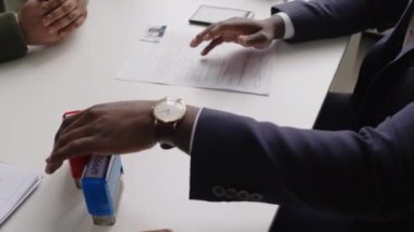 Siyahi erkek konsolosluk görevlisinin, elçilikteki görüşme sırasında başvuranların vize belgelerine reddedilen damgayı yapıştırdığı yüksek açılı bir fotoğraf.