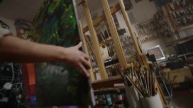 Orta yaşlı beyaz erkek sanatçı, kıvırcık saçlı ve sakallı, resim sehpasına renkli ve soyut bir resim koyarak işe hazırlanıyor.