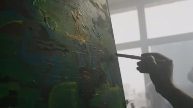 Avangard resim sanatına detaylar ve renk aksanları ekleyerek, puslu bir günde, stüdyoda resim sehpasında çalışırken, tanınamayan erkek sanatçının yakın plan çekimleri.