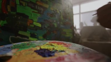Parlak akrilik ya da yağlı boya lekeleri olan paletin yakın çekimi, ve arka planda soyut başyapıt üzerinde çalışan anonim ressamın eli, pencereden gelen bulanık ışık