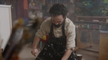 Kafkasyalı, önlüklü, stüdyoda oturmuş, kahverengi el işi kağıdına sarılı bir teli boyayıp alıcıya göndermeye hazırlanan yetişkin bir erkek sanatçının orta derece eğim yavaşlığı.