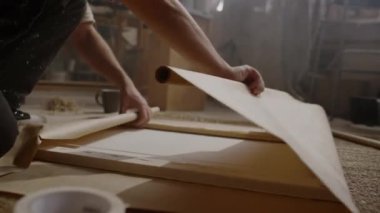 Anonim erkek ressamın el ve ayaklarının yakın plan çekimi. Atölyede yerde oturuyor. Tabloyu alıcıya göndermek için el işi kağıdına sarıyorlar.
