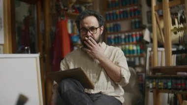 Orta yaşlı, beyaz sakallı, gözlüklü bir erkek sanatçının atölyede sandalyeye oturmuş, ekranda olmayan natürmort çalışırken ve not defterinde çizim yaparken,