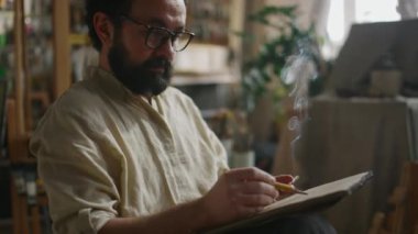 Olgun sakallı beyaz erkek ressamın stüdyoda otururken, sigara içerek sigara içerek resim ve sanat eserlerini çizdiği orta ölçekli bir resim.