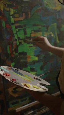 Parlak aksan renkleri ve fırçasıyla paleti tutan, parlak avantgarde yağı veya akrilik resim üzerinde çalışan tanınmayan erkek sanatçının orta boy dikey omuz üstü çekimi, küçük ayrıntılar ekliyor.