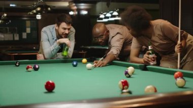 Kafkasyalı erkek ve siyahi kadının bilardo oynarken arkadaşlarının bilardo topuna vuruşunu ve bilardo oynarken bilardo topunu deliğe batırışını izlediği orta boy bir fotoğraf.