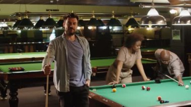 Akşamları çeşitli arkadaşlarla oynarken bilardo kulübünde bilardo masasında bilardo sopasıyla poz veren sakallı beyaz adamın orta boy portresi. Kameraya bakın.
