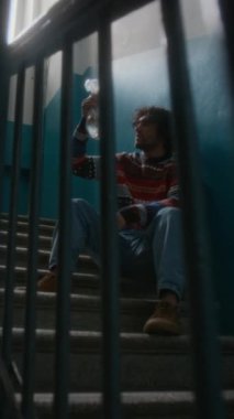 Vertical full lowkey shot of lonely man in sweater and jeans sitting on steps in stairway landing, looking at pet fish in clear bag and questioning meaning of life