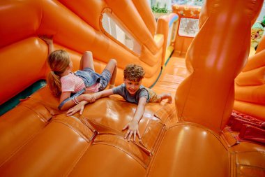 Mutlu küçük oğlan ve sarışın kız sarp, yumuşak turuncu deri kaydırağın üzerinde yatarken yuvarlanmamaya çalışıyorlar.